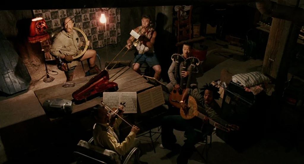 The Ladykillers (2004) movie still: wide — A cluttered, dimly lit basement contains musical equipment and a makeshift rehearsal area. Four diverse…