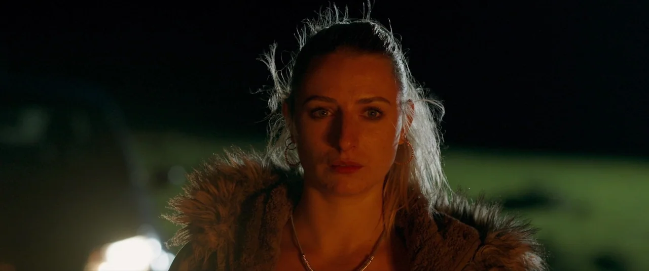 Bull (2021) movie still: closeup — An open field at night is illuminated by a singular bright vehicle light. A woman stands in the beam of light…