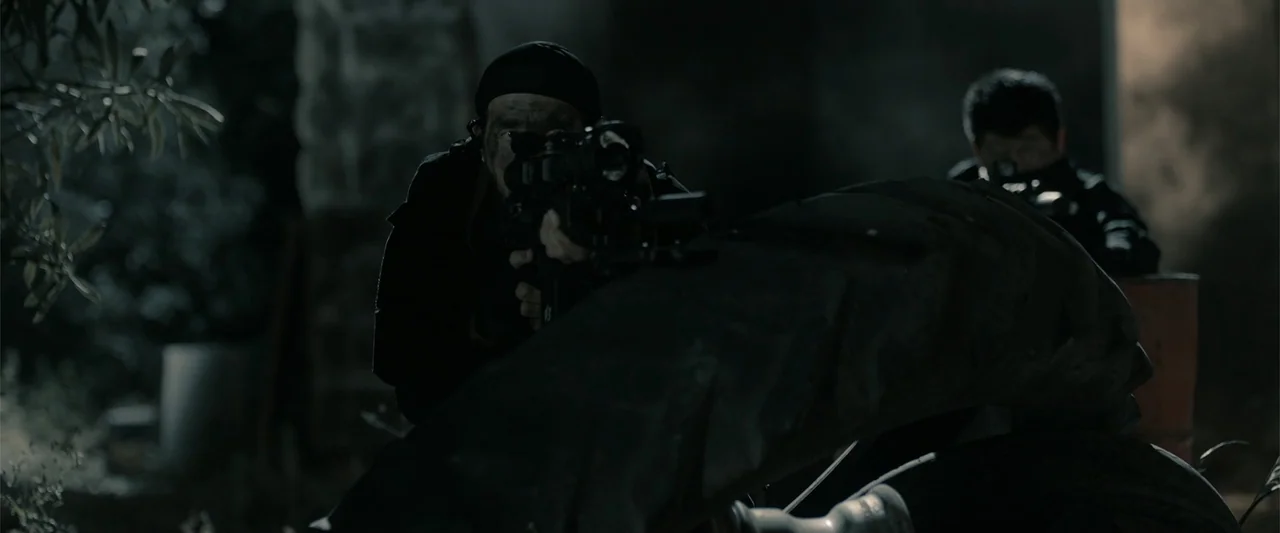Hounds of War (2024) movie still: medium — A dark, rubble-strewn outdoor area at night with heavy shadows and low visibility. Two men in tactical…