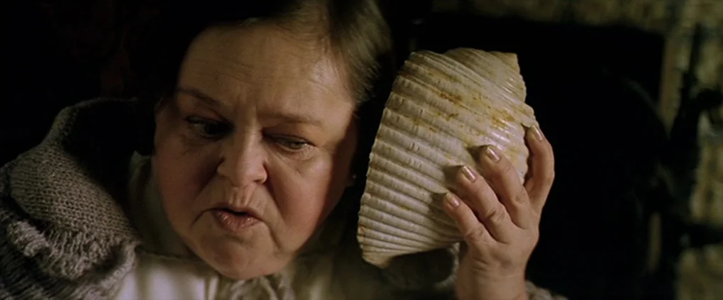 Anguish (1987) movie still: closeup — A dimly lit room with muted earth tones and a textured wall backdrop. An older woman holds a large seashell…