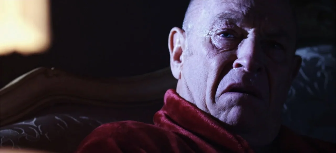 The Russian Bride (2019) movie still: closeup — A dimly lit room at night with a plush armchair and shadowed background. An elderly man in a red…