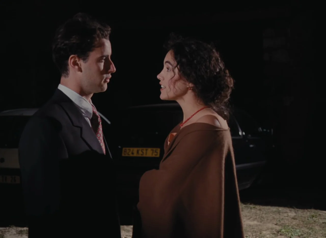Autumn Tale (1998) movie still: medium — Two individuals stand in an outdoor space at night near parked vehicles. The man in a suit faces the woman…