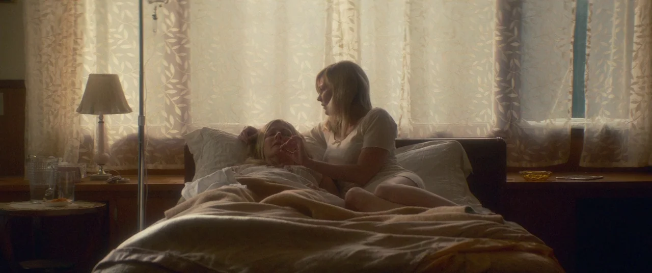 Woodshock (2017) movie still: wide — A patient lies in a hospital bed beside a window with patterned sheer curtains. A woman in a white shirt sits…