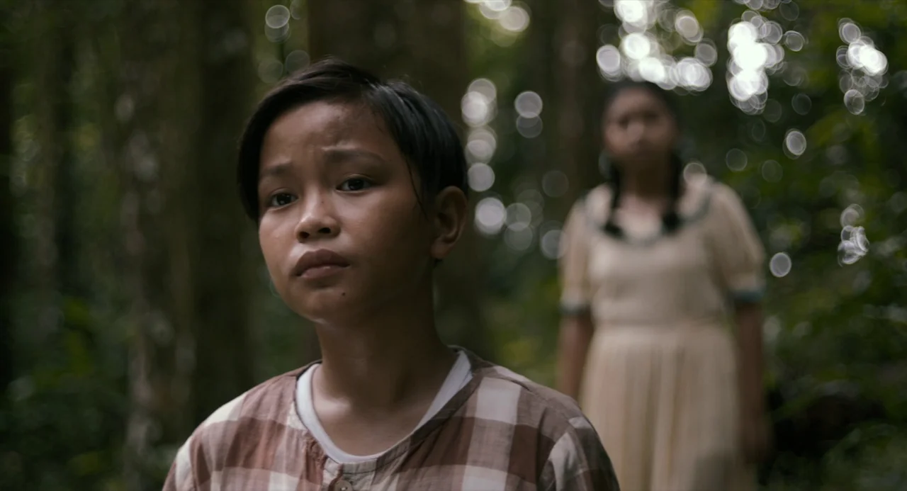 In My Mother’s Skin (2023) movie still: medium — A dense forest environment with out-of-focus bokeh highlights visible among the deep foliage. A…