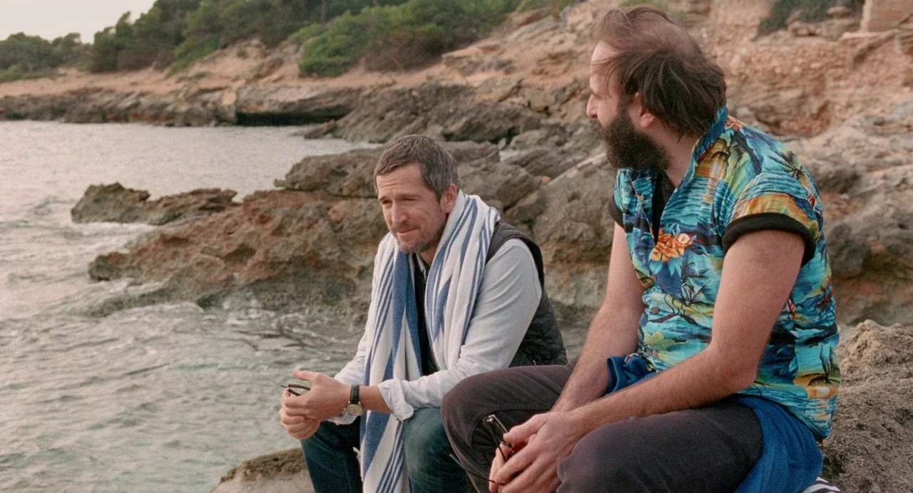 Non-Fiction (2018) movie still: medium — A rocky shoreline sits beside calm coastal water with sparse vegetation on the slopes. Two men sit on the…