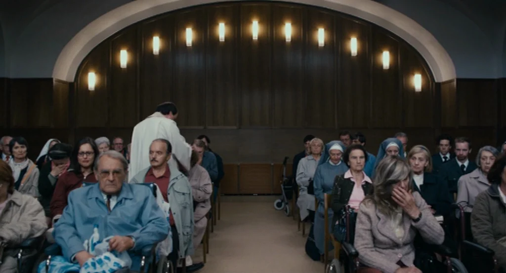 Lourdes movie still: wide — A spacious hall features wood-paneled walls accented by a series of vertical wall sconces. Rows of people are seated in…