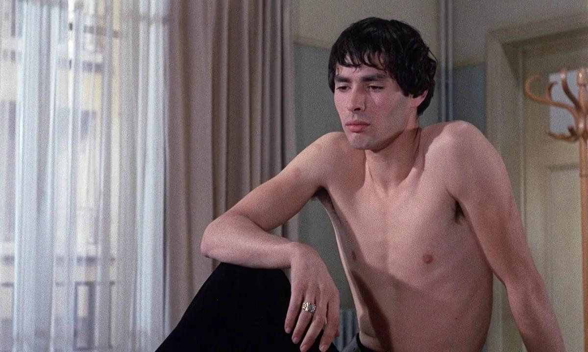 Belle De Jour (1967) movie still: medium — A room with light-colored walls and sheer white curtains covering a large window. A shirtless man sits in…
