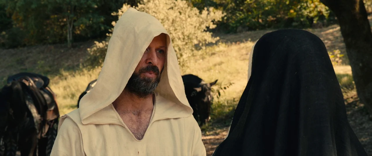 Benedetta (2021) movie still: medium — An outdoor trail with light foliage and horses stands in the background. A bearded man in a hooded tunic…