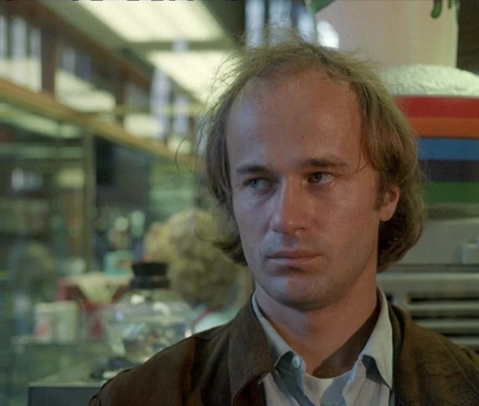 Crime & Punishment (1983) movie still: closeup — A man stands inside a 1970s retail shop with merchandise blurred in the background. He looks past…