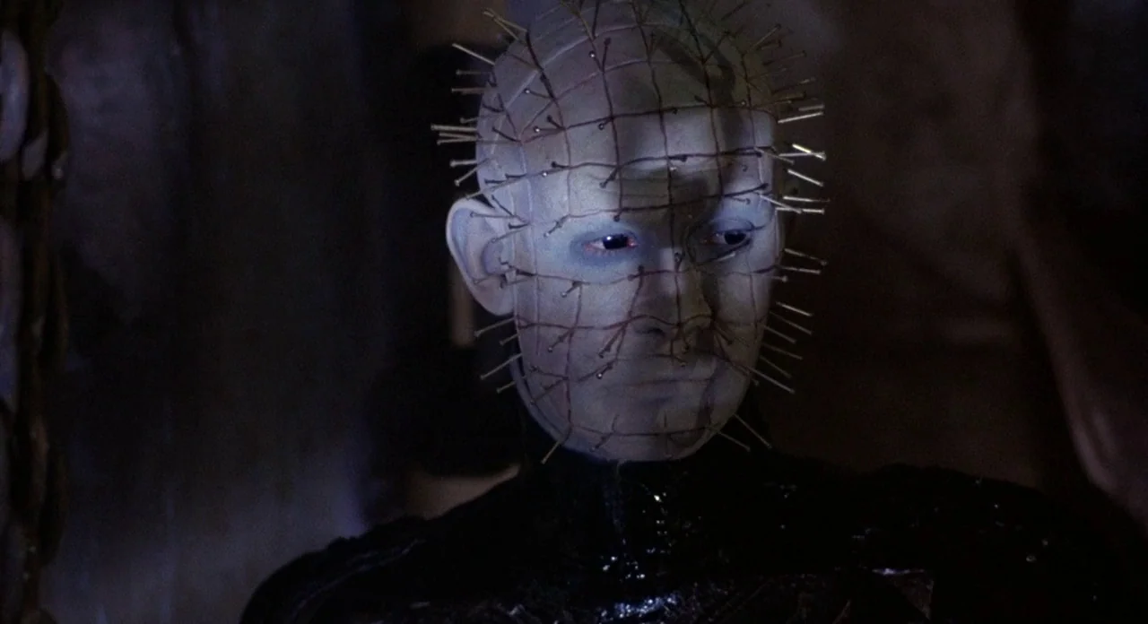 Hellraiser (1987) movie still: closeup — A dark, nondescript interior space with minimal architectural detail visible in the shadows. A pale,…