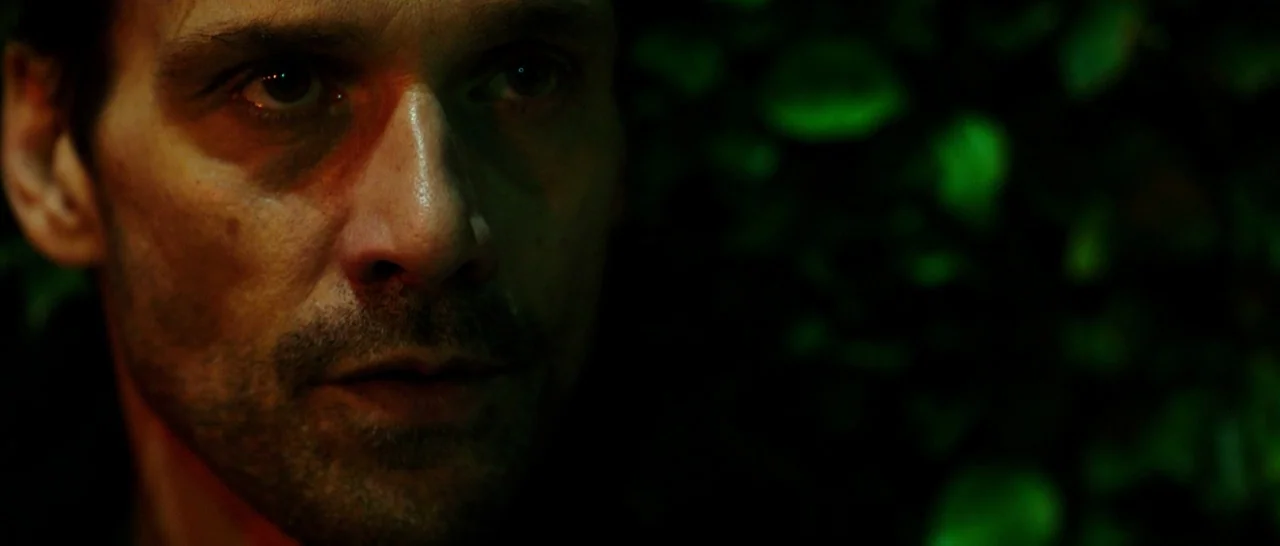 The Purge: Anarchy (2014) movie still: closeup — A dark outdoor setting featuring dense green foliage visible behind a man. The man stares directly…