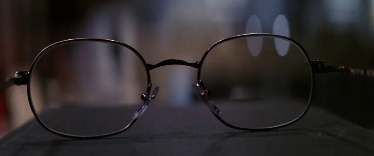 Mission: Impossible (1996) movie still: closeup — A pair of thin metal-rimmed spectacles sits on a dark, matte surface. The background is a soft,…