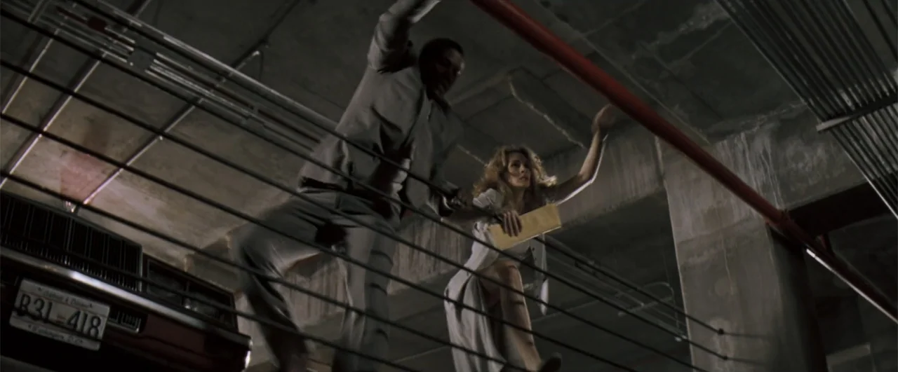 The Pelican Brief (1993) movie still: fullbody — A concrete parking garage with exposed overhead piping and a multi-level wire security barrier. A…