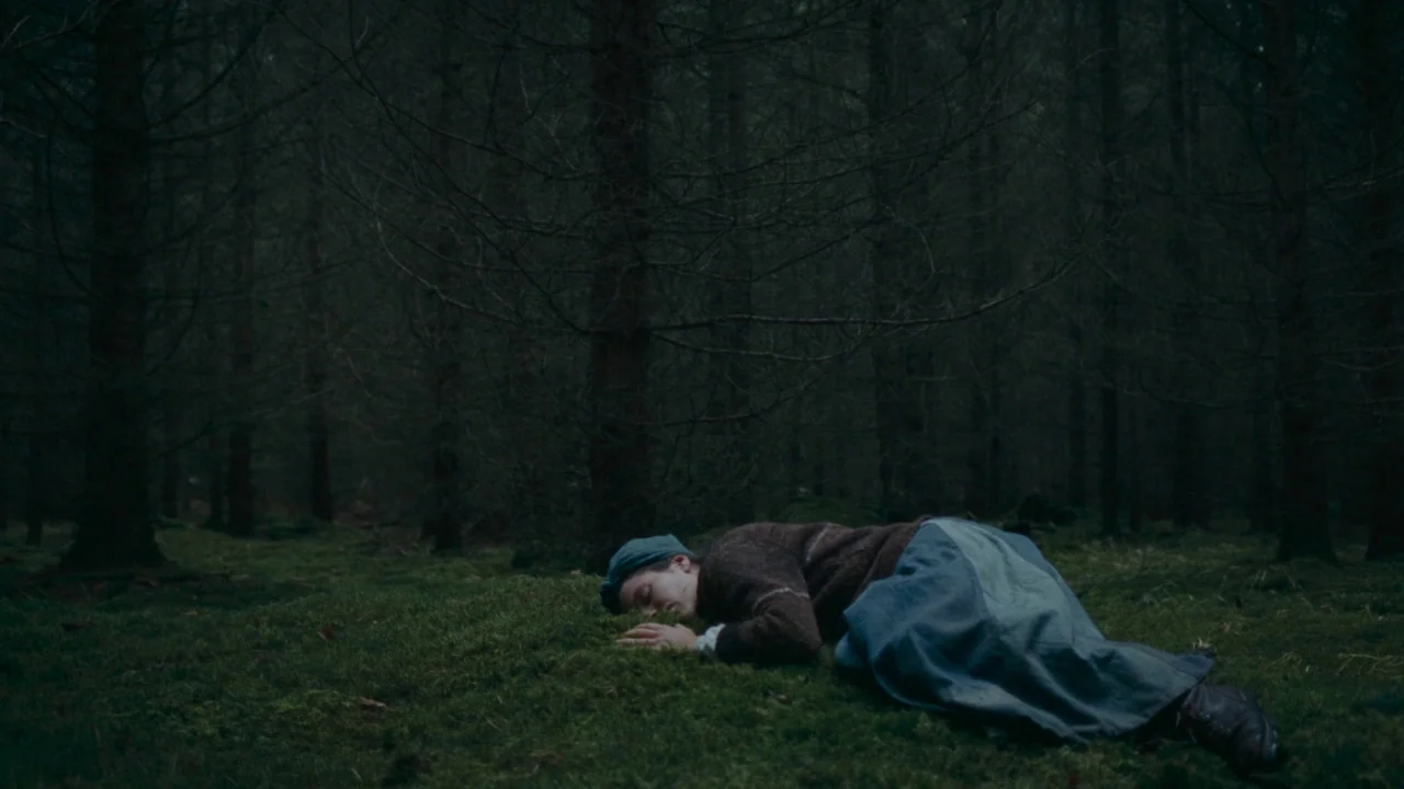 The Devil’s Bath (2024) movie still: wide — A dense coniferous forest features a mossy ground layer under tall, thin dark trees. A young woman lies…