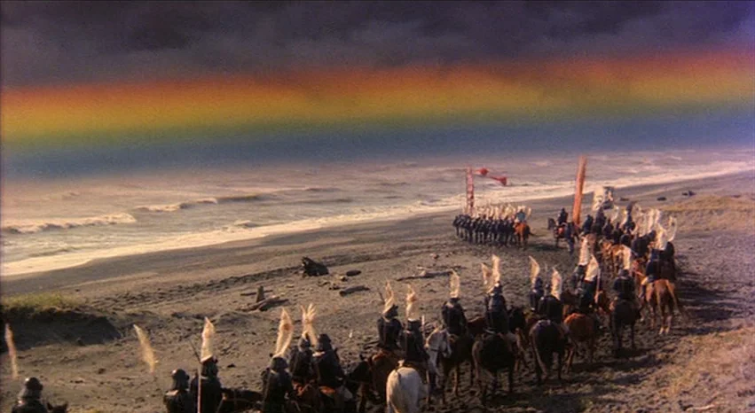Kagemusha (1980) movie still: wide — A long line of samurai warriors on horses rides along a dark, empty beach. A prominent rainbow stretches…
