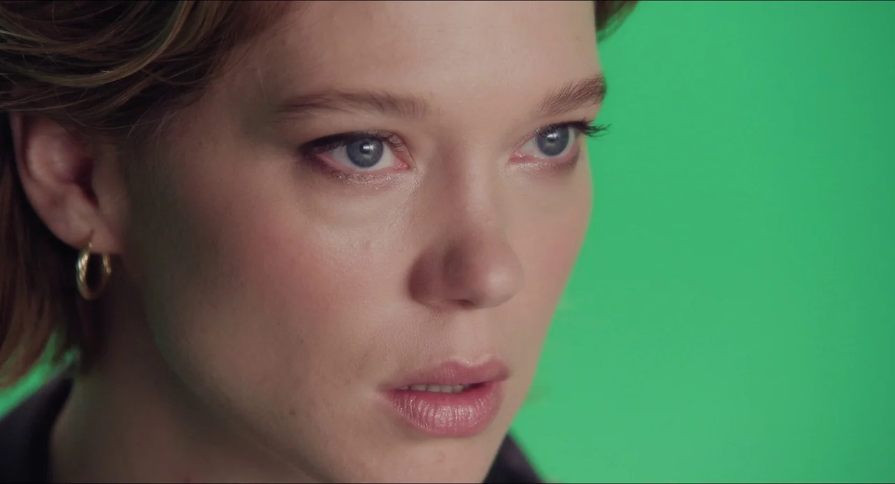 Transformers: Rise of the Beasts (2023) movie still: closeup — A woman stands in front of a solid green screen backdrop. The frame captures her face…