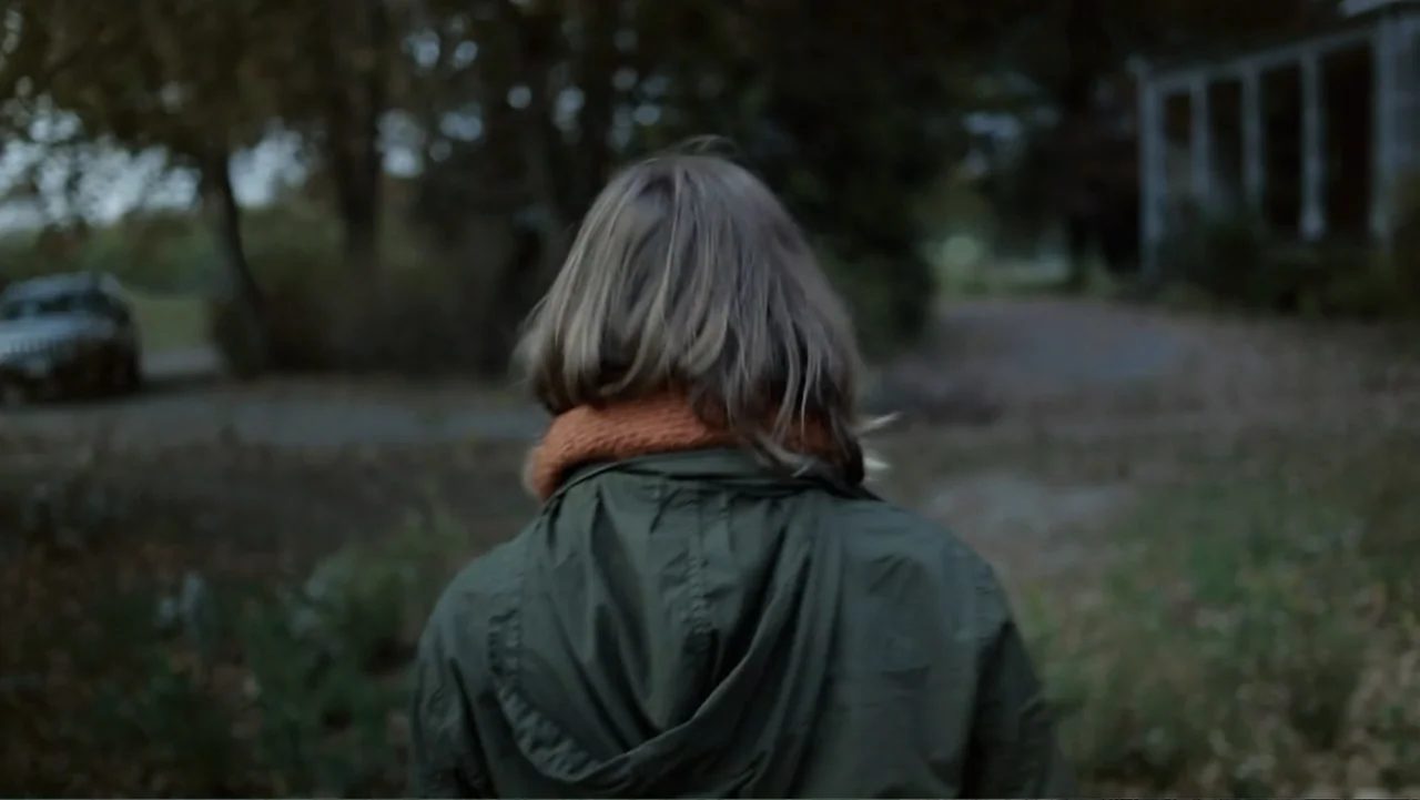 Silent House (2011) movie still: medium — A rural outdoor setting with a gravel driveway leading toward a blurred house facade. A person with…