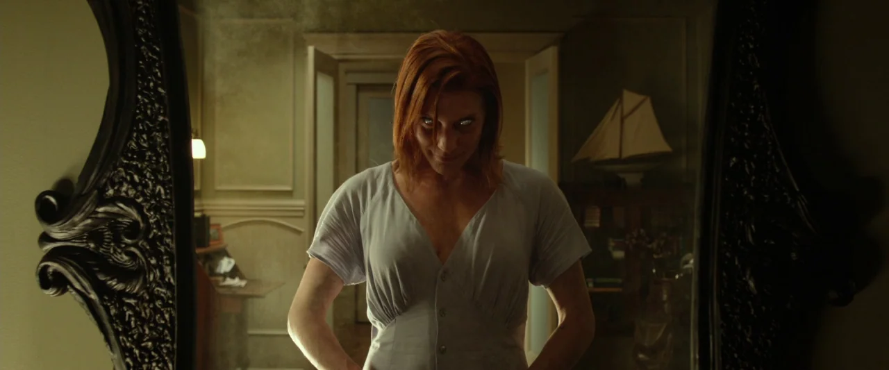 Oculus (2013) movie still: medium — An interior bedroom features a large dark, ornate mirror reflecting a woman standing before it. A model ship…