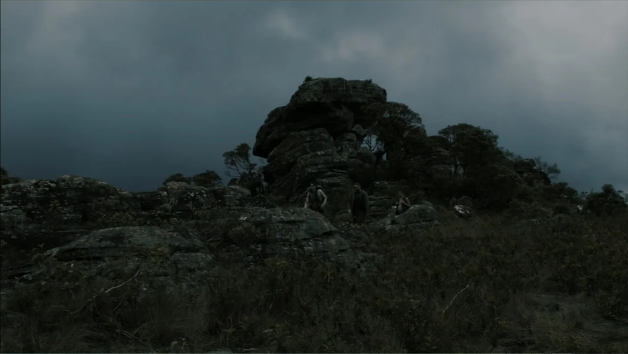 The Grotto (2022) movie still: wide — A rugged landscape features large weathered rock formations surrounded by low brush under clouds. Three people…