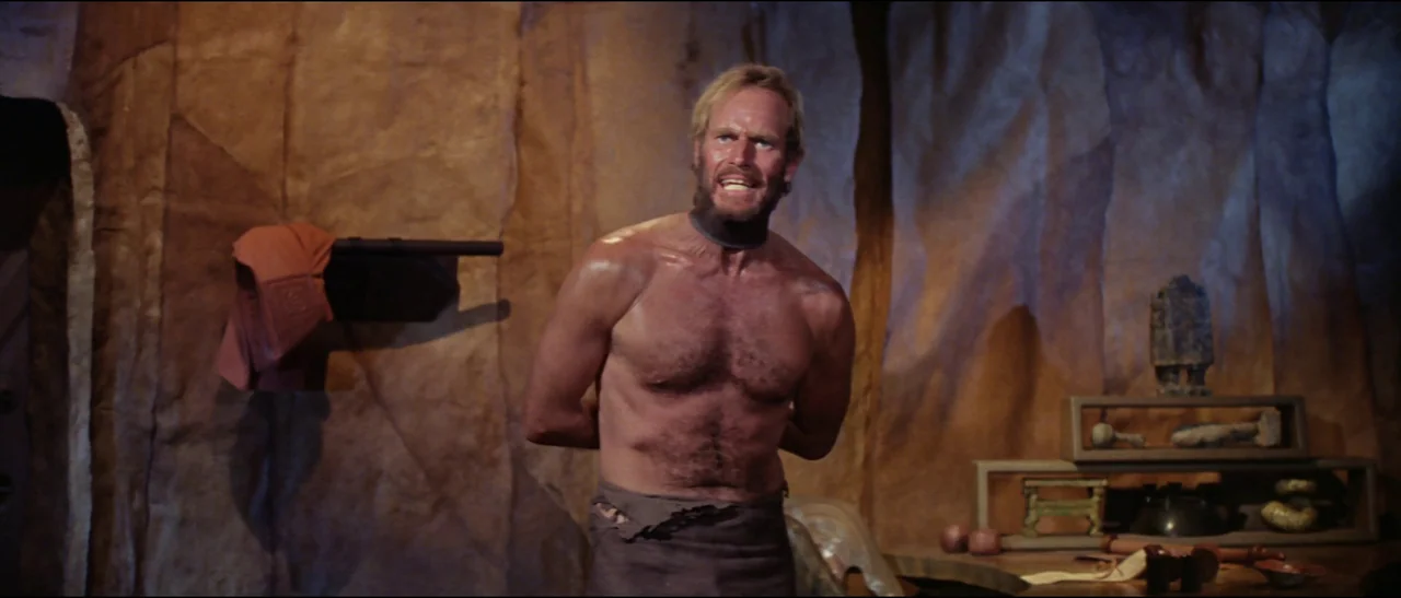 Planet of the Apes (1968) (1968) movie still: medium — A dimly lit stone cave with textured rock walls and primitive decorative objects. A muscular…