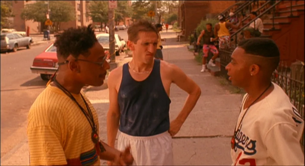 Do The Right Thing (1989) movie still: medium — A sunny Brooklyn city block features brownstone buildings and parked cars along the curb. Three…