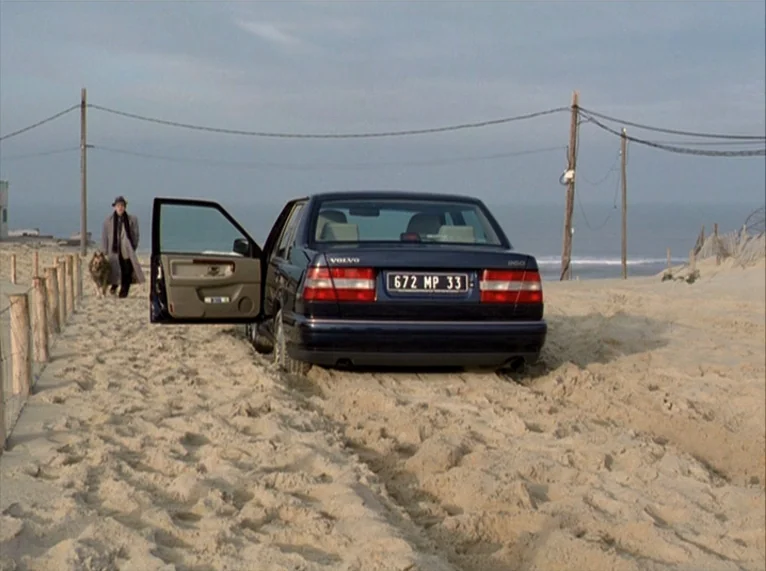 For Ever Mozart (1996) movie still: wide — A dark blue sedan is parked on a sandy beach under an overcast sky. An older man in an overcoat walks…