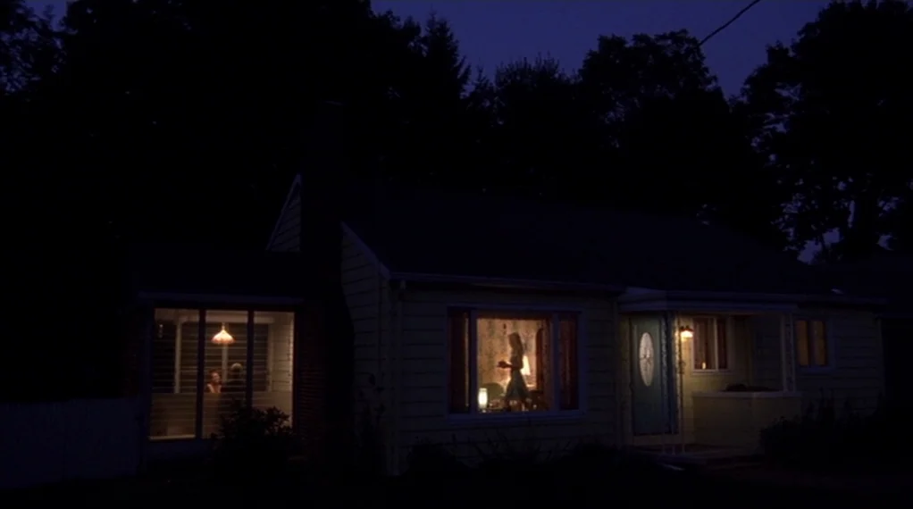 Broken Flowers movie still: wide — A modest yellow one-story house stands under a dark night sky. Two illuminated windows reveal silhouettes of…