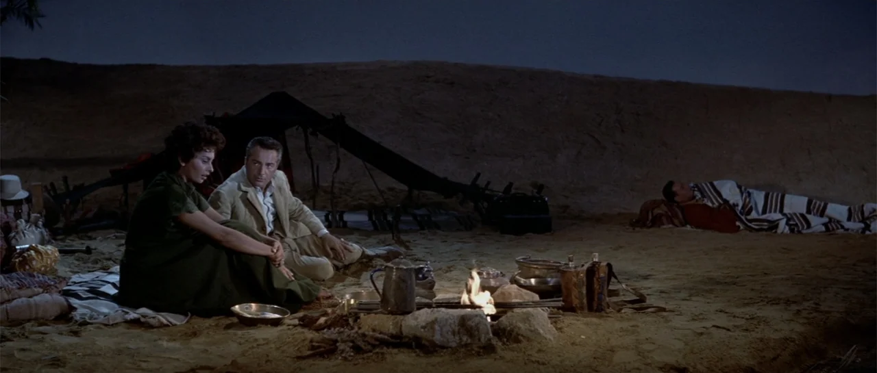 Legend of the Lost (1957) movie still: wide — A sandy desert landscape at night features a small fire and scattered camping gear. Two people sit…