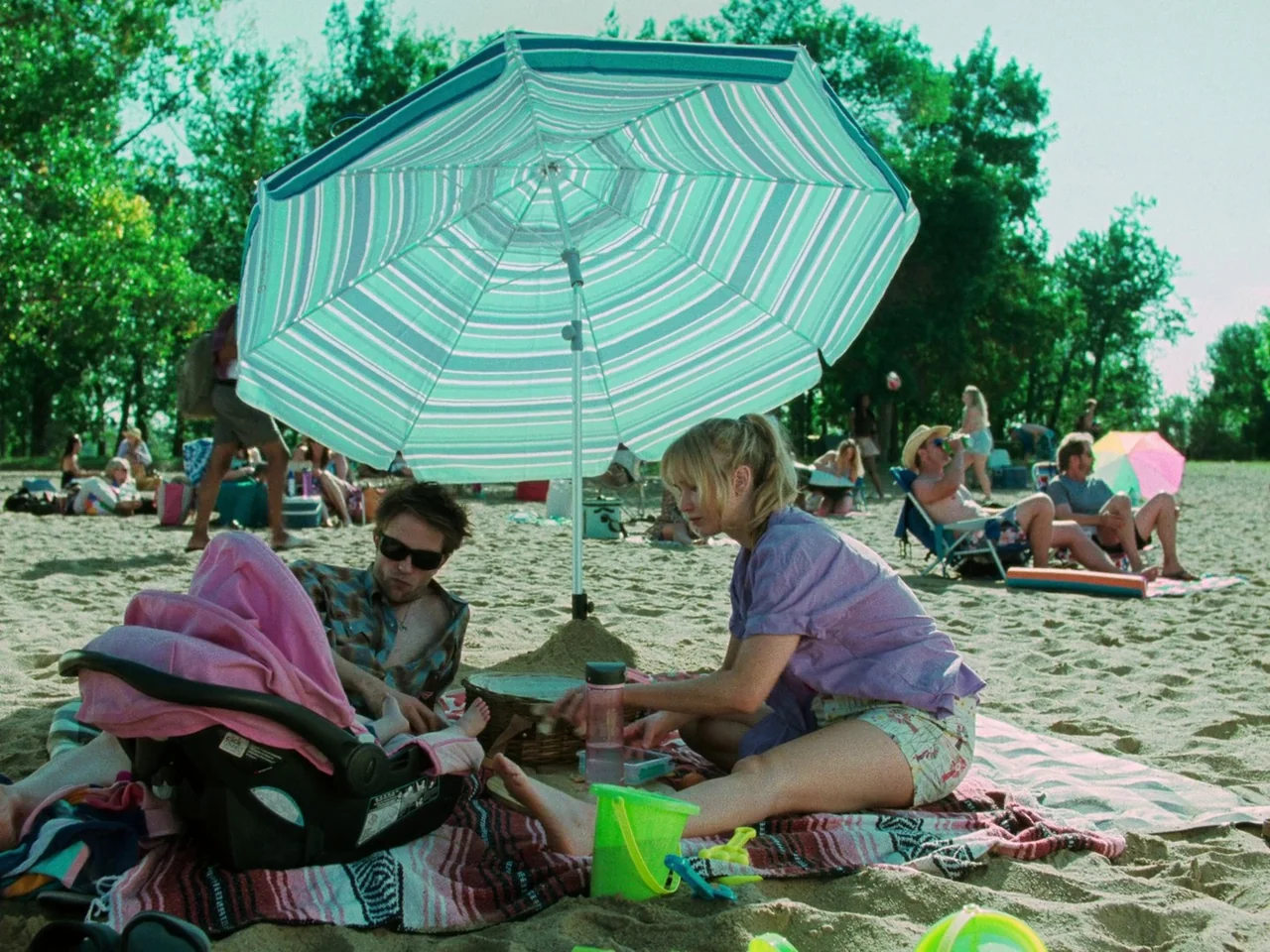 Die My Love (2025) movie still: medium — A sunny public beach filled with people sits under a canopy of trees. A couple sits on a blanket beneath a…