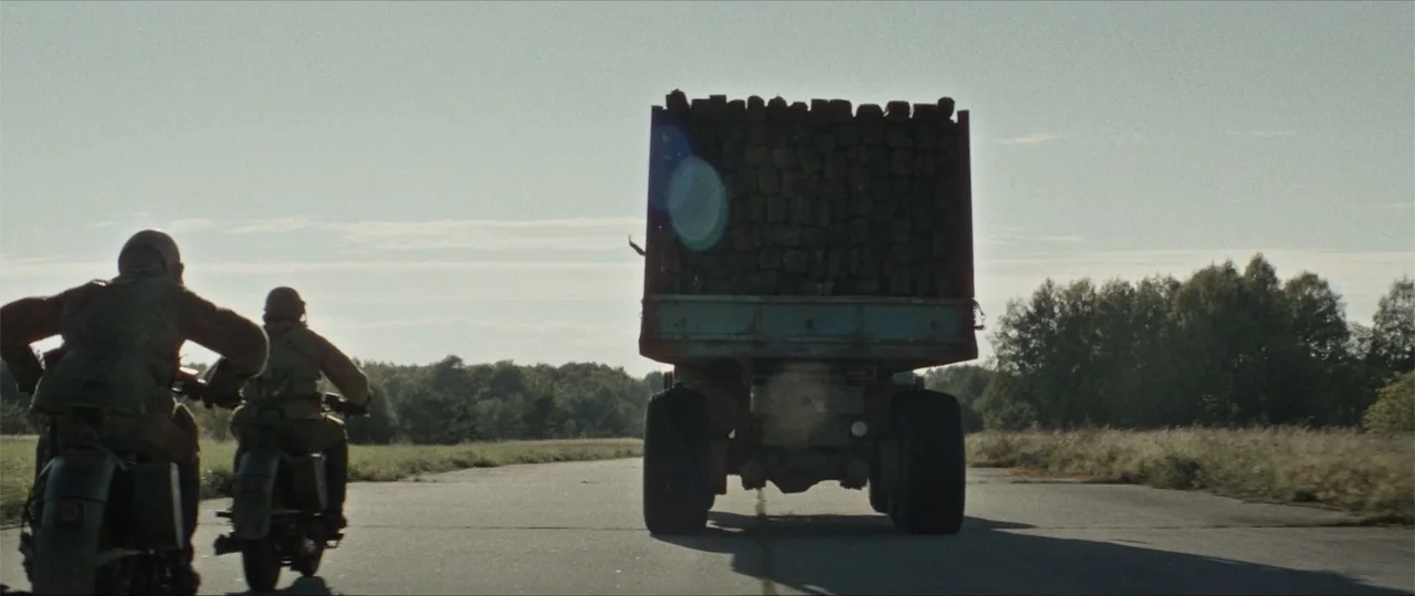 Sisu: Road to Revenge (2025) movie still: wide — An asphalt road stretches across an open landscape during a bright, overcast day. Two soldiers on…