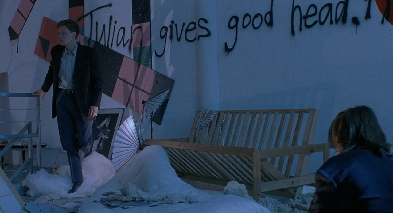 Less Than Zero (1987) movie still: wide — A sparse room features a wooden futon frame and graffiti sprayed across a wall. A man stands amidst…