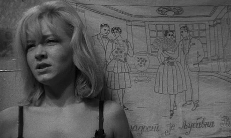 Man Is Not Bird (1965) movie still: closeup — A woman stands before a wall hanging featuring line-drawn illustrations of dancing figures. The…