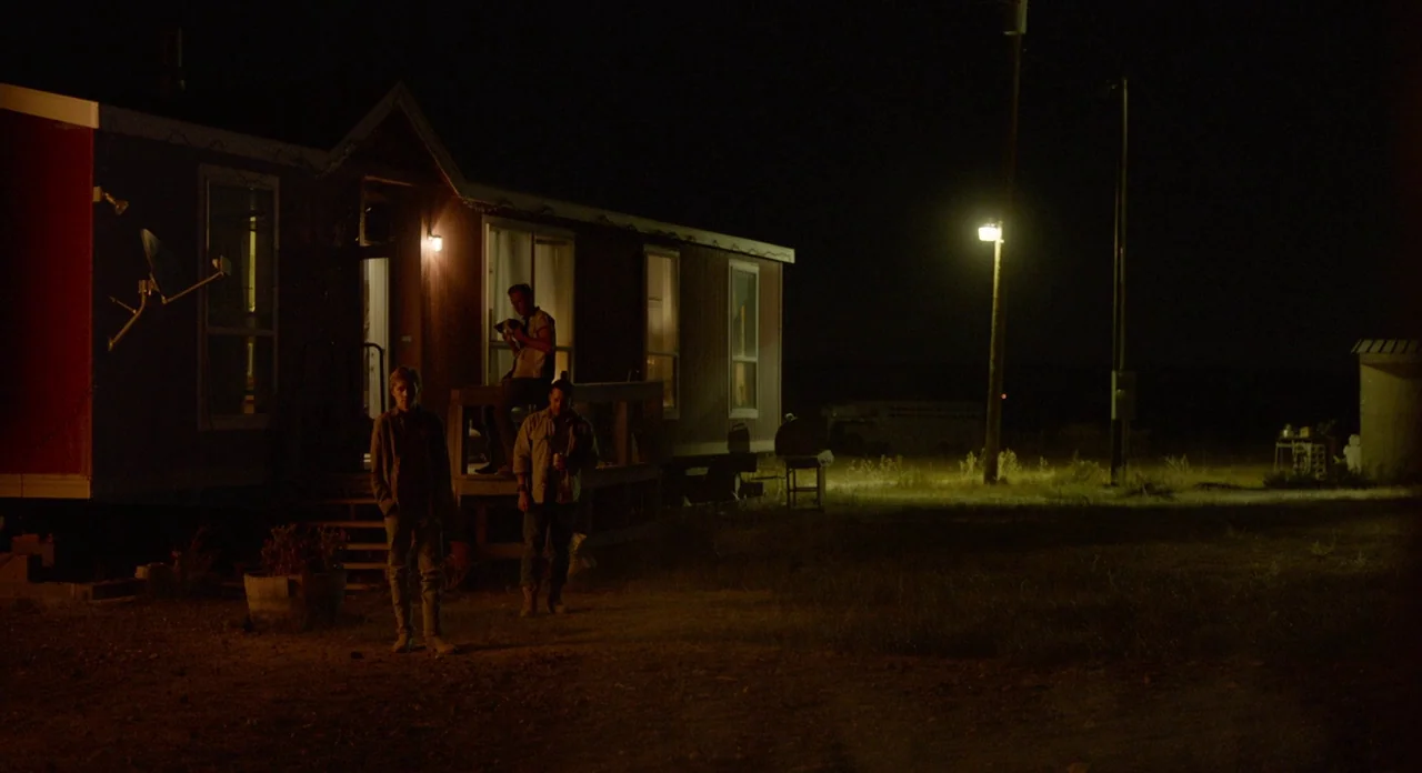 Lean on Pete (2017) movie still: wide — A reddish mobile home sits in a dark, rural field at night. Three men stand near the porch steps while a…