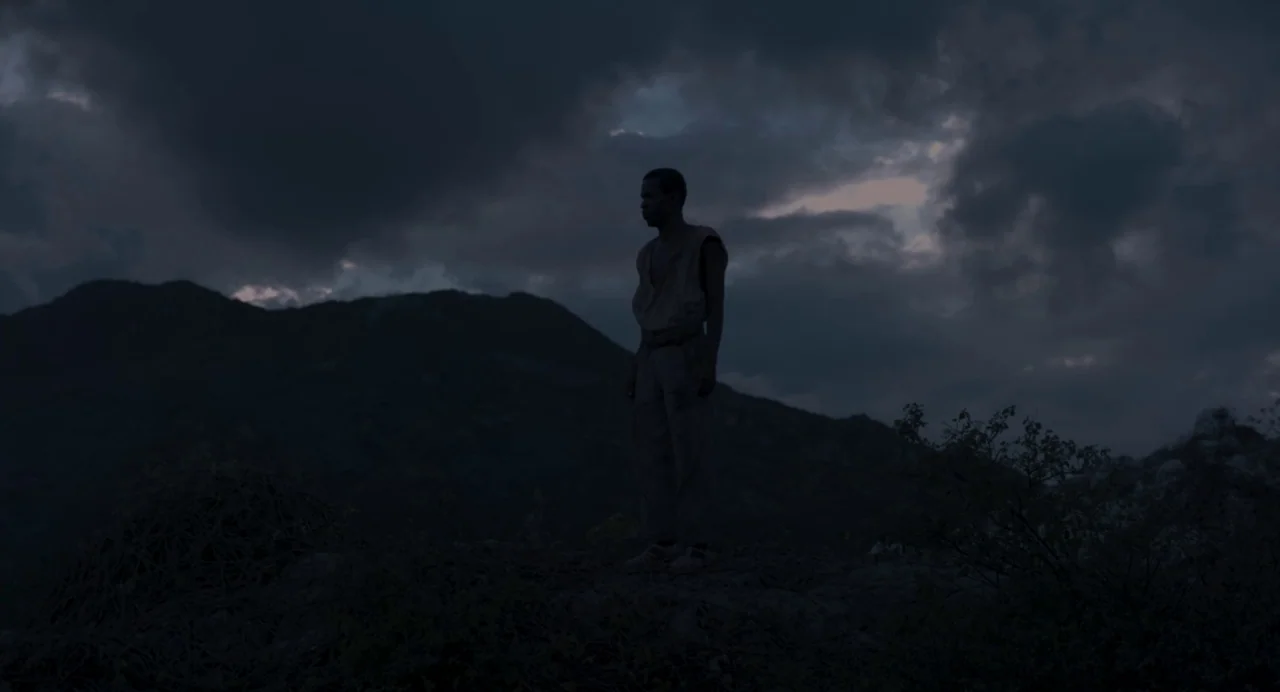 Zombi Child (2019) movie still: fullbody — A dark mountainous landscape with heavy clouds fills the evening sky. A man stands silhouetted on a rocky…