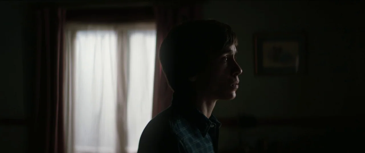 Good Favour (2017) movie still: medium — A dimly lit room with a window covered by translucent curtains at the back. A young man stands in the dark…