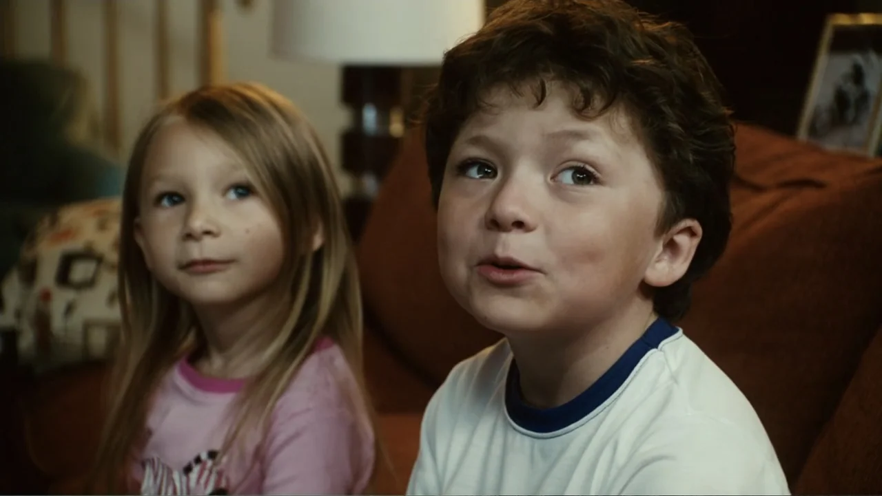 Click (2006) movie still: medium — A domestic living room features a dark fabric sofa in the background. A young boy in a white shirt sits next to a…
