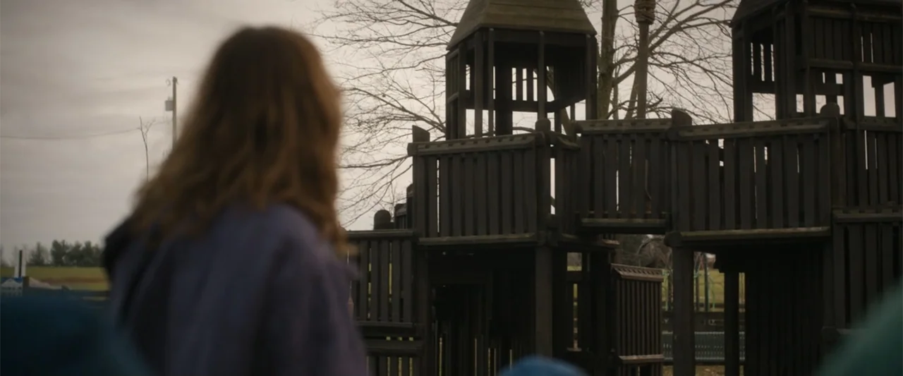 Unsung Hero (2024) movie still: medium — A wooden play structure stands in a desolate area under a cloudy sky. A person stands in the foreground…