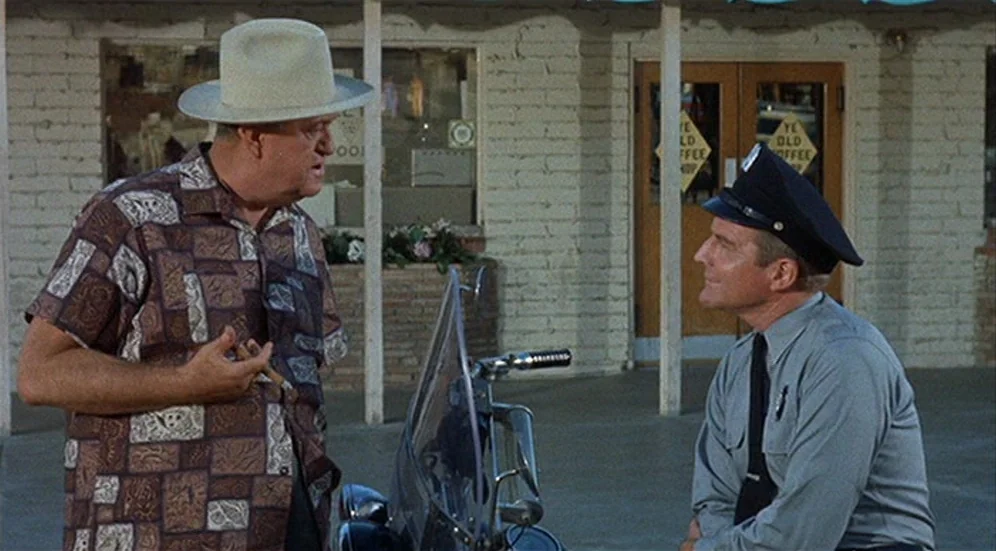 Kid Galahad (1962) movie still: medium — An older man in a hat talks to a police officer outside a building. The storefront has a white brick facade…