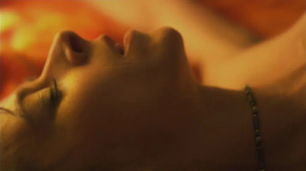 Red Road (2006) movie still: closeup — A dark, warm-toned interior space with soft, directional lighting. A woman lies on her back with her head…
