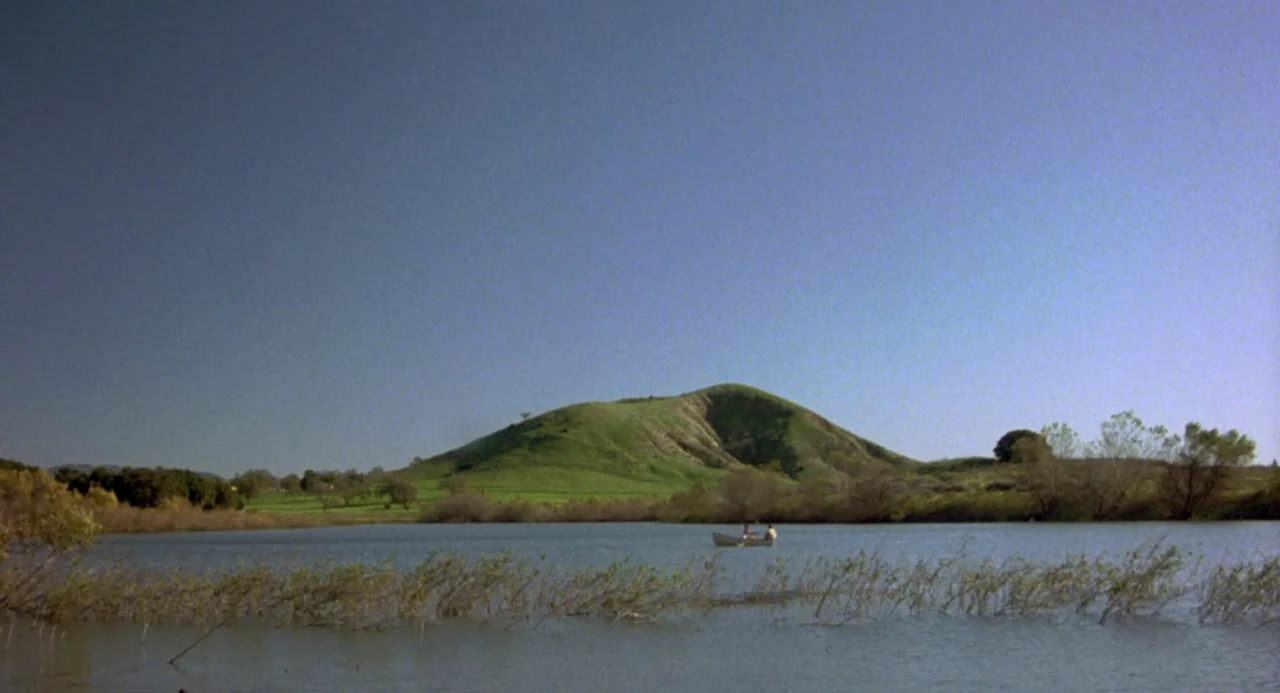 The Karate Kid (1984) movie still: establishing — A calm lake sits before a large, grassy, rounded hill under a clear sky. A tiny white rowboat with…