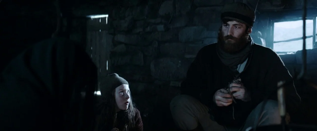 Black ’47 (2018) movie still: medium — A rustic stone-walled room is lit only by faint natural light from a window. An bearded man and a young child…