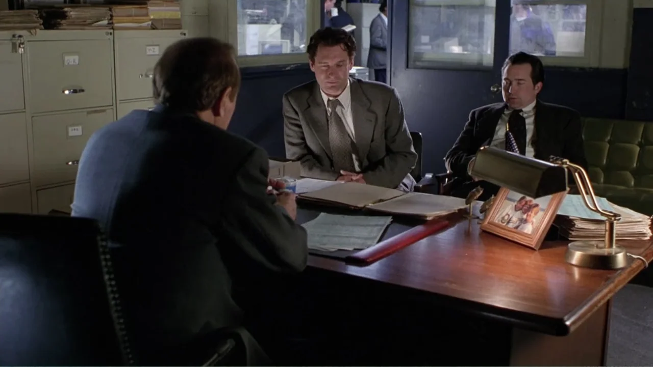 Mistrial (1996) movie still: medium — A dimly lit office features metal filing cabinets and a large wooden desk. Three men in suits conduct a…