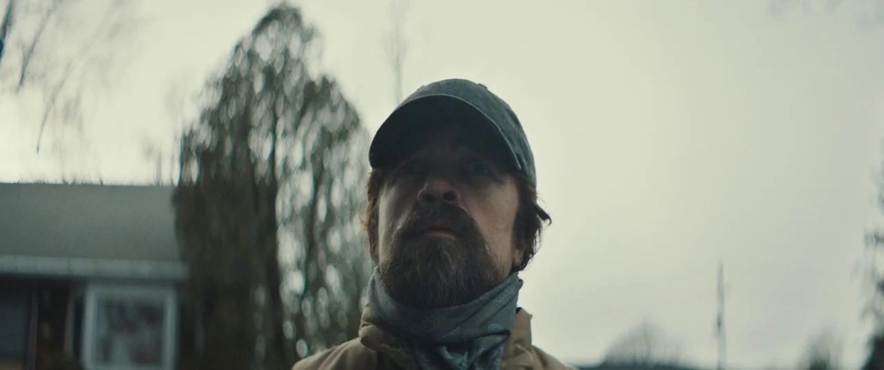 I Think We’re Alone Now (2018) movie still: closeup — A residential street under an overcast sky with bare trees and a distant house. A man wearing…