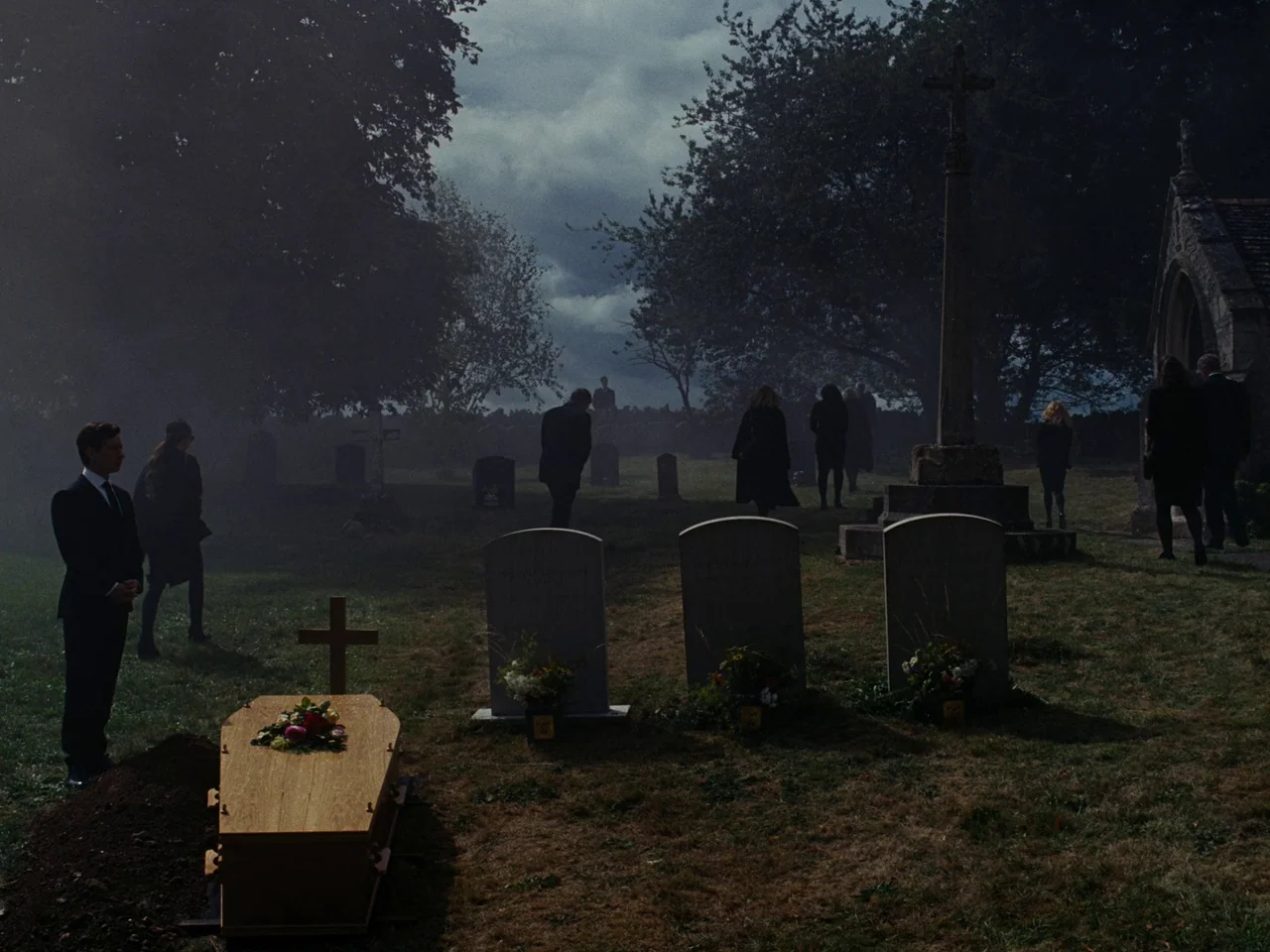 Saltburn (2023) movie still: wide — A group of people in dark clothing gathers in an overcast outdoor cemetery. A light-wood coffin sits in the…
