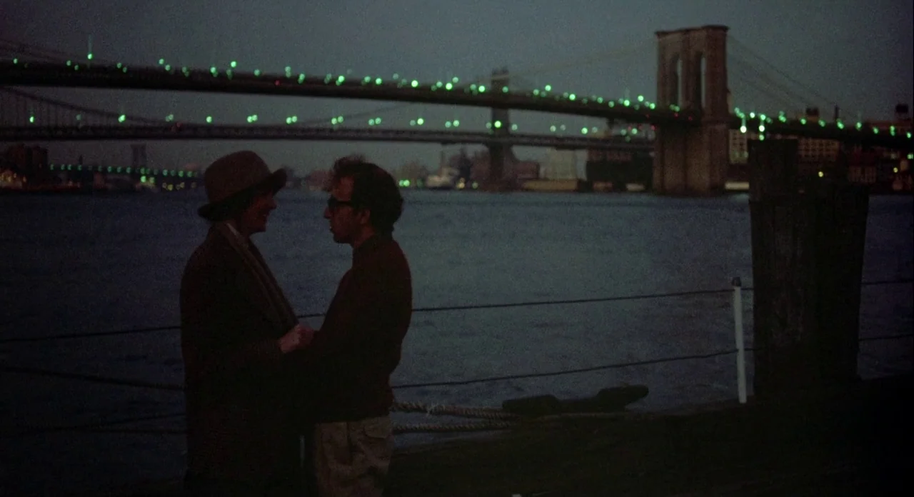 Annie Hall (1977) movie still: fullbody — A dark riverfront at night with the lit Brooklyn Bridge spanning the background. A man and a woman stand…