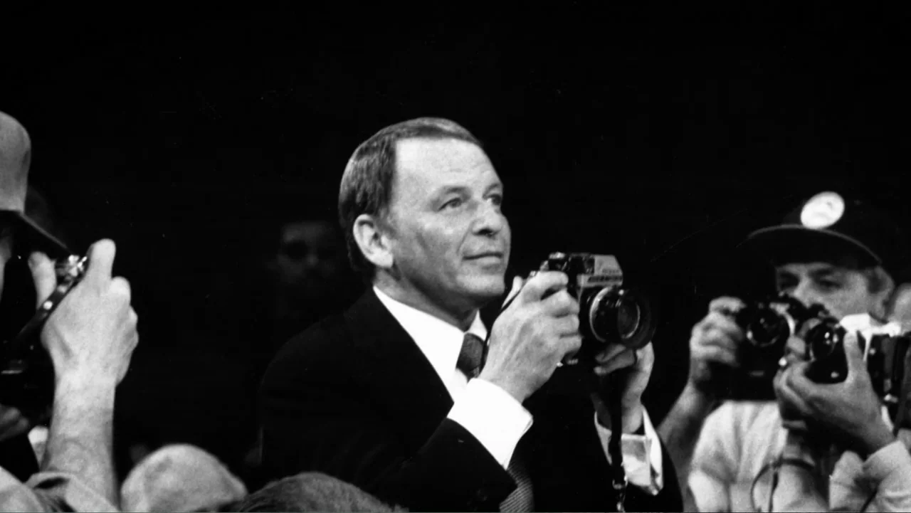 Ali & Cavett: The Tale of the Tapes (2018) movie still: medium — A man in a suit stands among a group of photographers in a dark room. The man holds…
