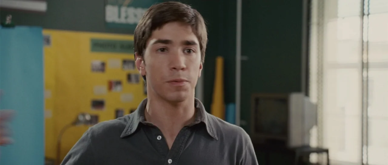 Accepted (2006) movie still: medium — A classroom interior with a bulletin board and a television set in the background. A young man in a dark grey…