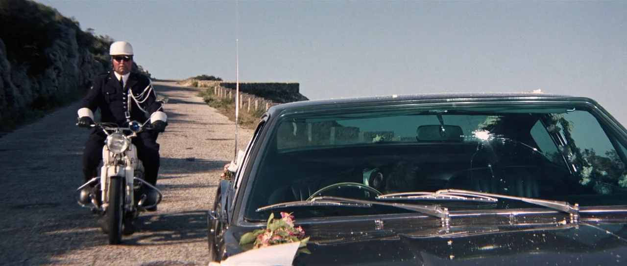 On Her Majesty’s Secret Service (1969) movie still: wide — A narrow stone road winds along a cliffside under a clear bright blue sky. A police…
