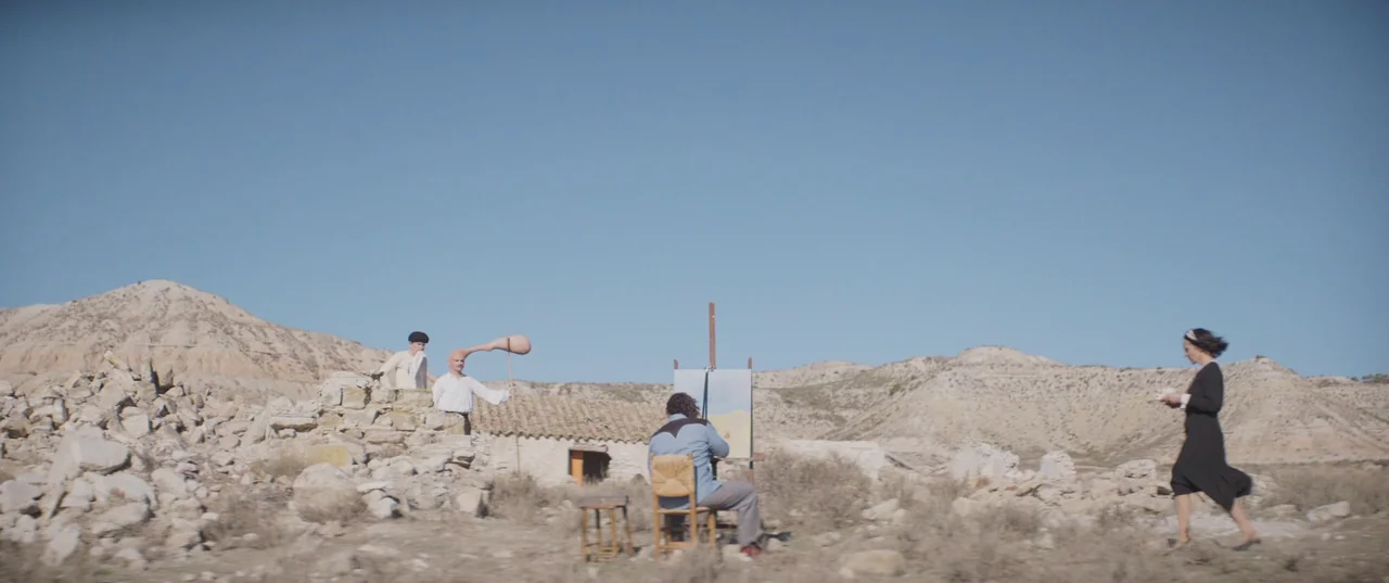 Daaaaaalí! (2023) movie still: wide — An arid, rocky desert landscape contains ruins of a stone building under blue sky. A man sits at an easel…