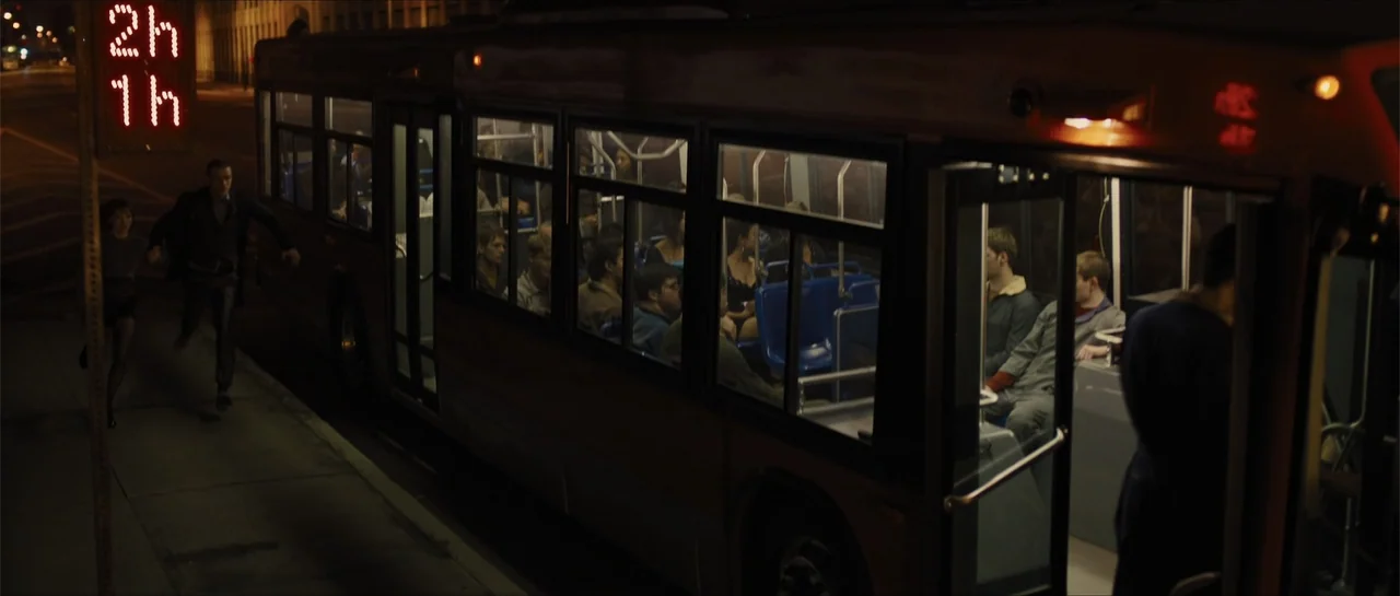 In Time (2011) movie still: wide — A city bus sits at a dark sidewalk stop at night under orange streetlights. A man in a dark blazer runs toward…